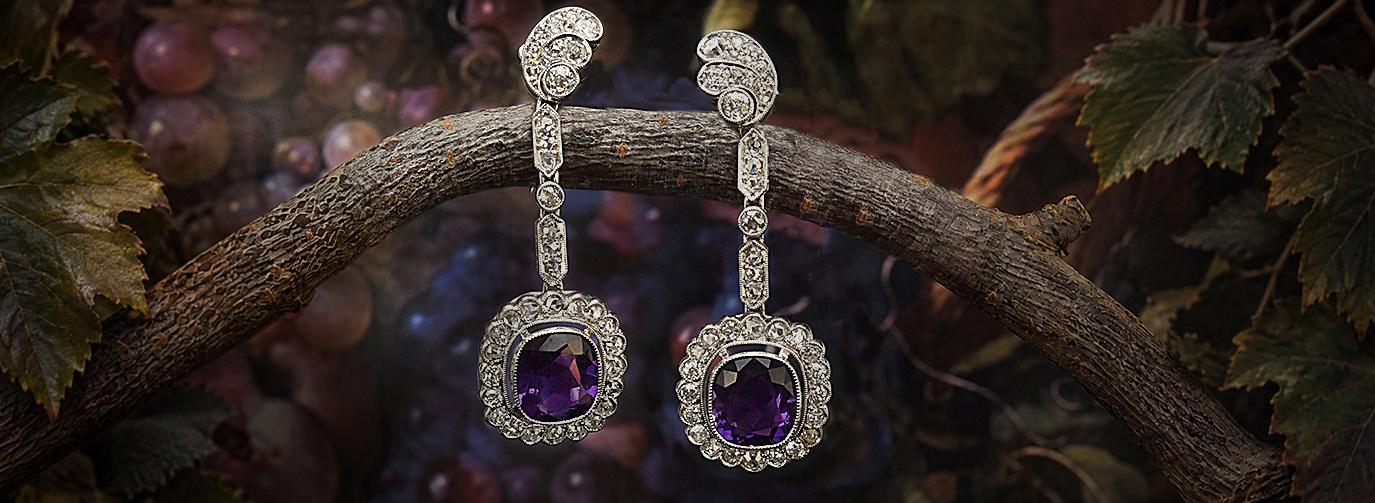 Ein paar lange Amethystohrringe mit Diamanten hängen über einen Ast. Im Hintergrund sind Weinblätter und Weintrauben zu sehen. 