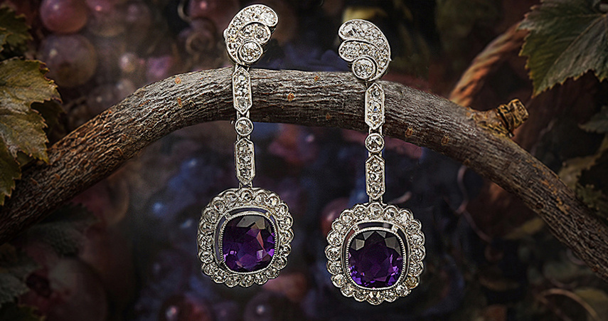 Ein paar lange Amethystohrringe mit Diamanten hängen über einen Ast. Im Hintergrund sind Weinblätter und Weintrauben zu sehen. 